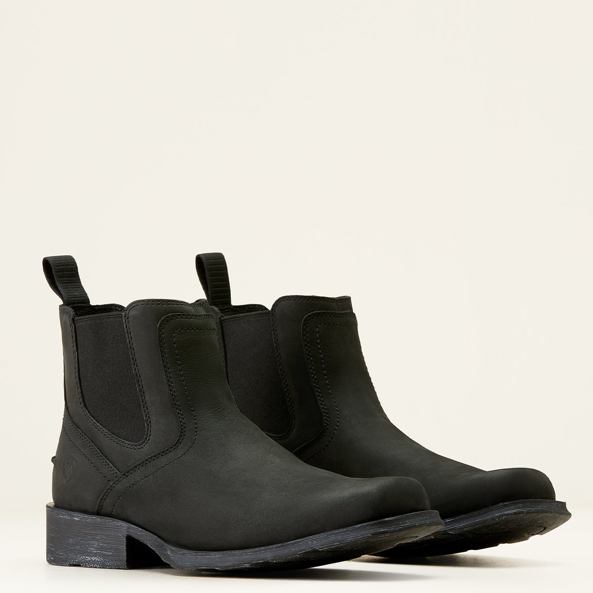 A pair of Ariat men's black Chelsea boots with elastic side panels and pull tabs, shown from a three-quarters angle on a light cream background.