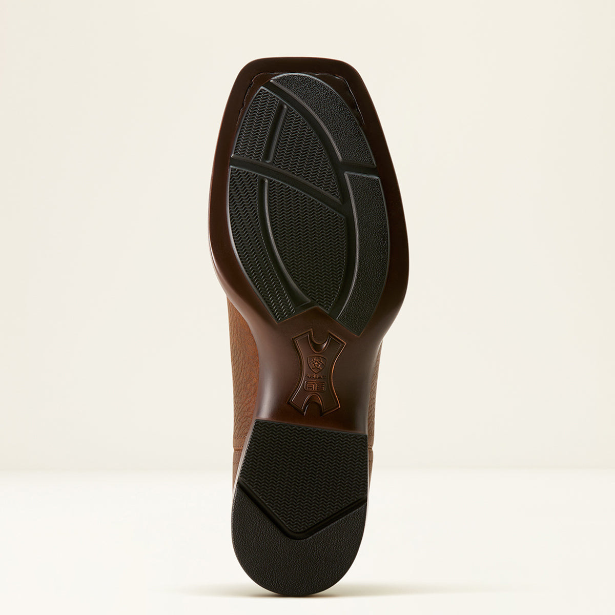 The Ariat outsole of a brown leather boot with a black rubber tread, shown from a top-down view against a light beige background.