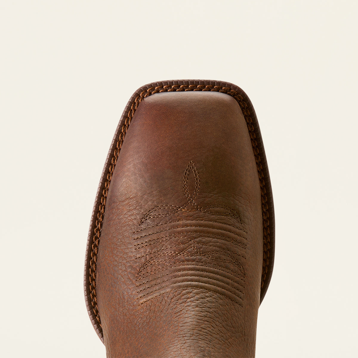 Close-up of the Ariat square toe of a brown leather cowboy boot with decorative stitching and a braided edge, shown against a light cream background.