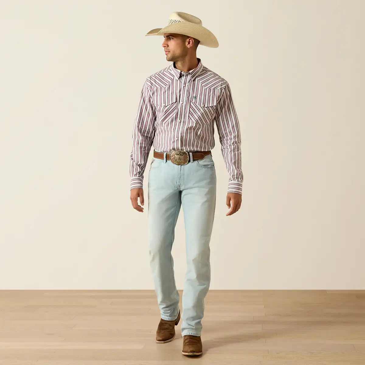 A man wearing a western outfit stands in profile against a plain beige studio background. He is wearing a light tan cowboy hat, a long-sleeve button-up Ariat shirt with vertical stripes in beige and reddish-brown, light-wash blue jeans, a brown belt with a large silver buckle, and brown cowboy boots.
