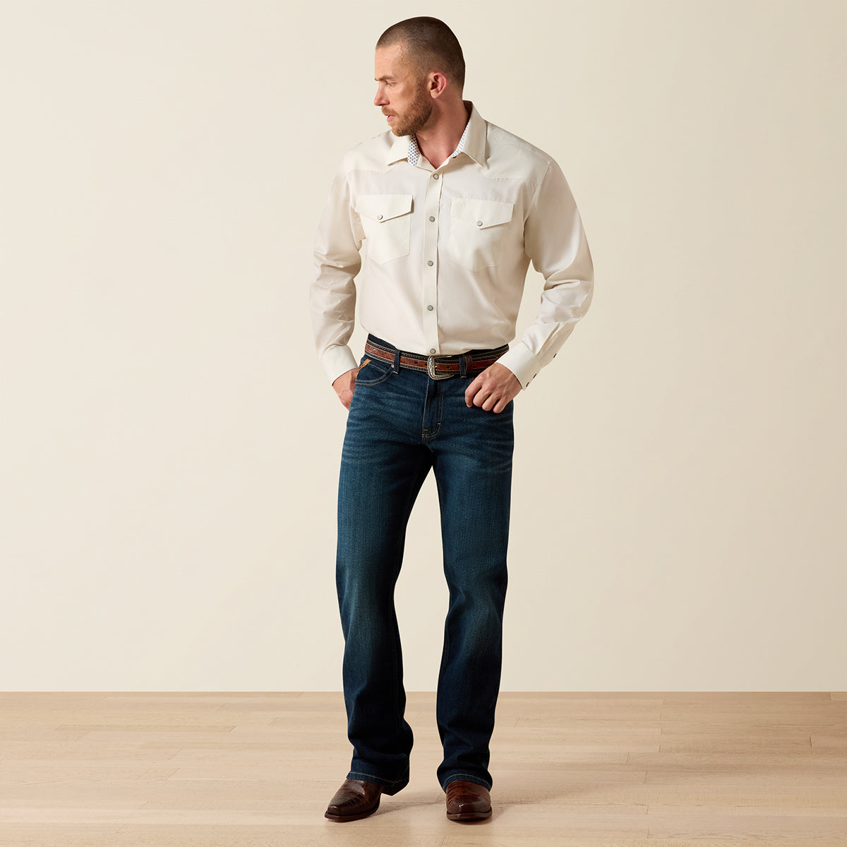 A man wearing a cream western-style button-down Ariat shirt and dark blue jeans, standing against a plain beige wall on a light wood floor.