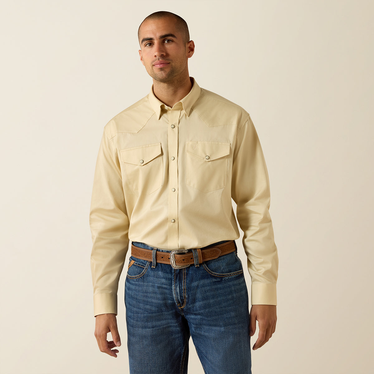 Man wearing a beige Ariat shirt and blue jeans against a plain background.