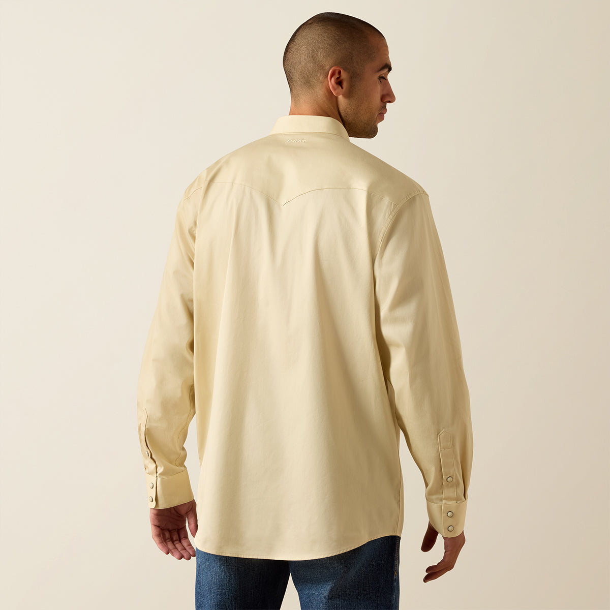 A man shown from the back wearing a light cream-colored long-sleeve button-up shirt and blue jeans, standing against a plain beige studio background.