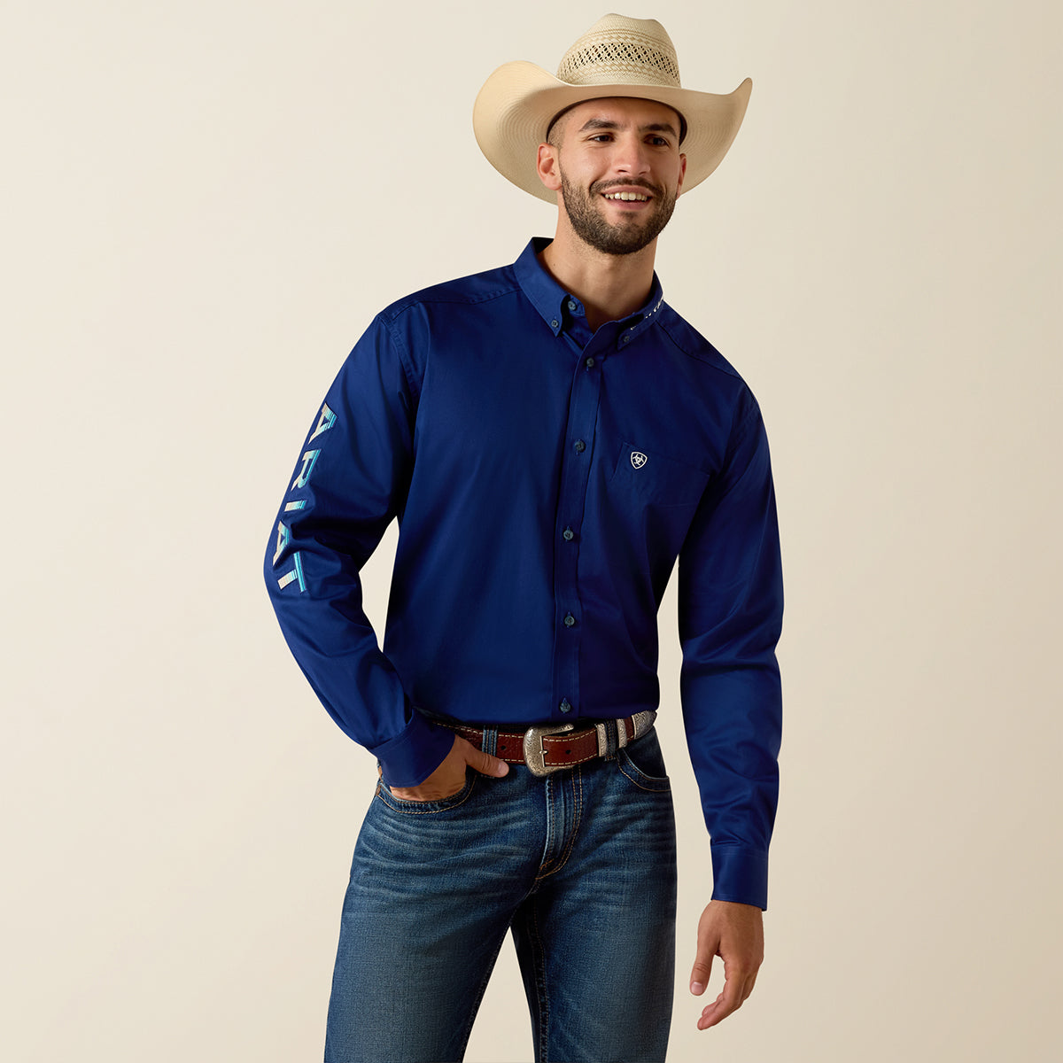 Man wearing a blue Ariat shirt with a logo, jeans, and a cowboy hat on a beige background