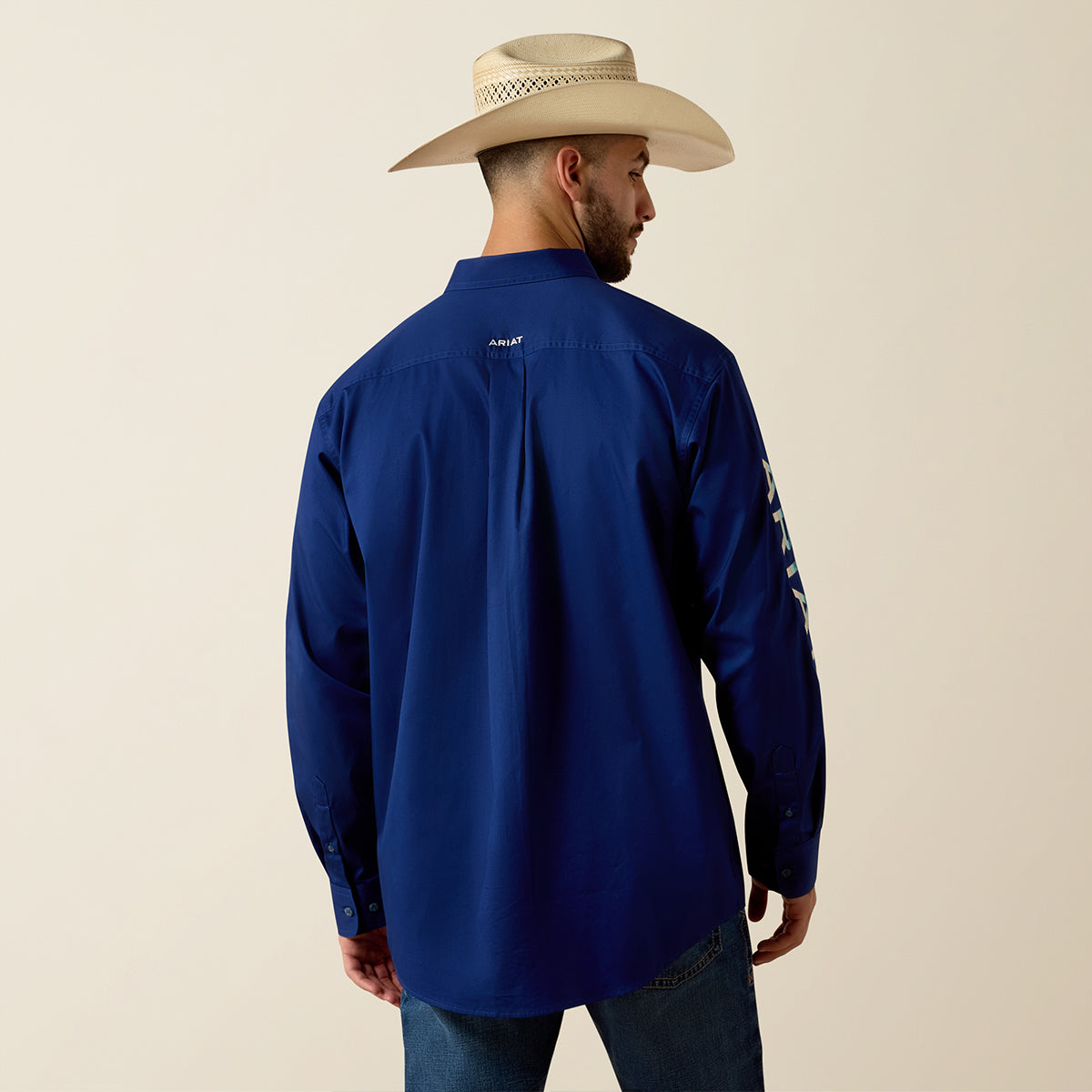 Man wearing a blue Ariat shirt with a brand logo on the back, standing against a beige background