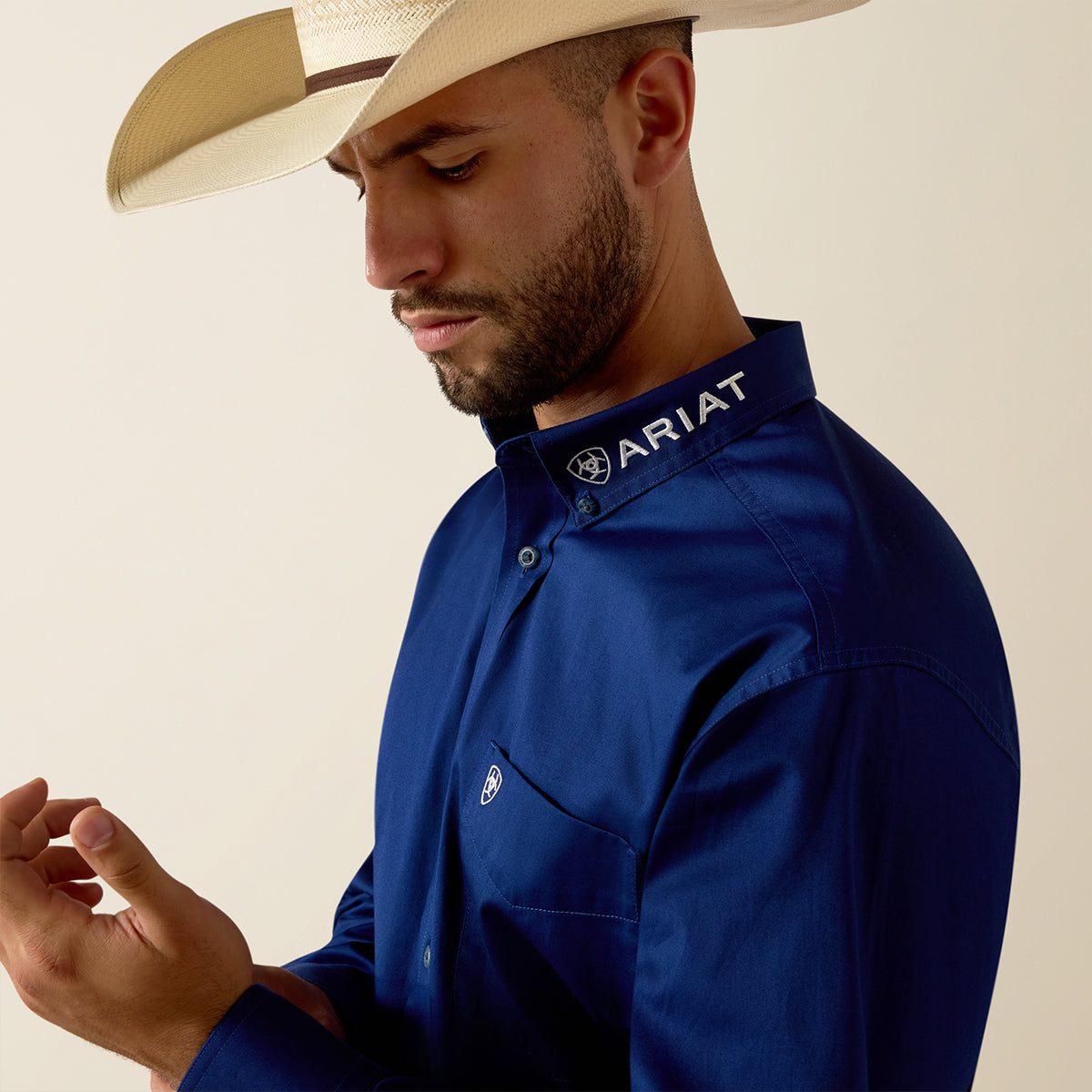 A man wearing a navy blue Ariat long-sleeve western shirt and a beige cowboy hat, shown in a close-up profile against a light beige background.