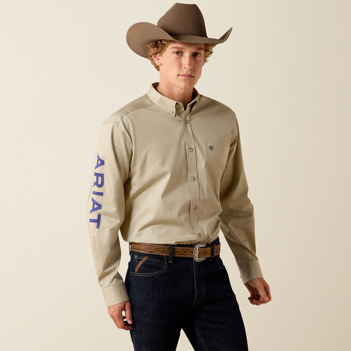 A man wearing a beige long-sleeve button-down shirt with 'ARIAT' printed in blue down the right sleeve, paired with dark jeans, a brown belt, and a brown cowboy hat, against a light beige studio background.