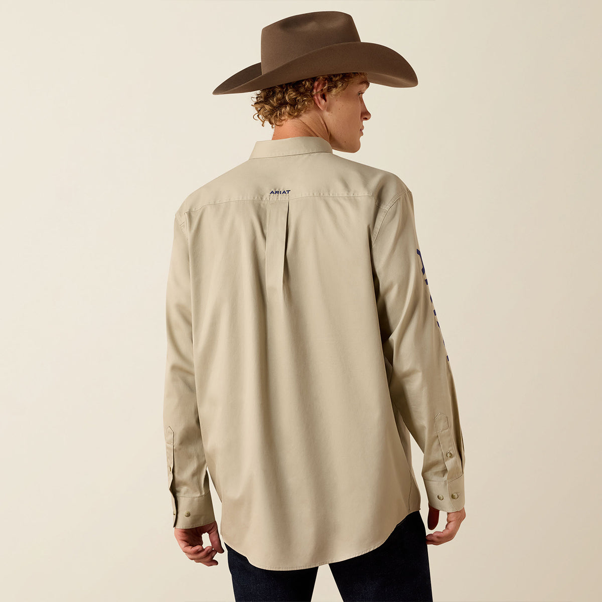 A man wearing a light beige western-style button-up shirt and a brown cowboy hat, photographed from the back against a plain light beige studio background.