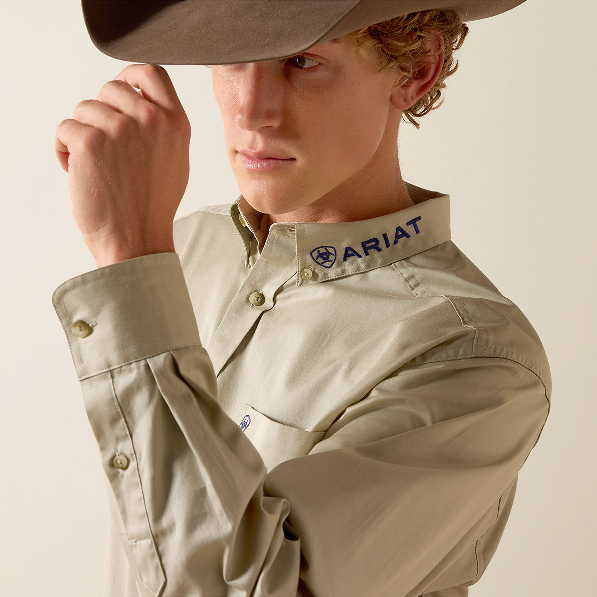 A close-up of a man wearing a light beige long-sleeve button-up shirt with the ARIAT logo on the collar, paired with a brown cowboy hat against a plain beige background.