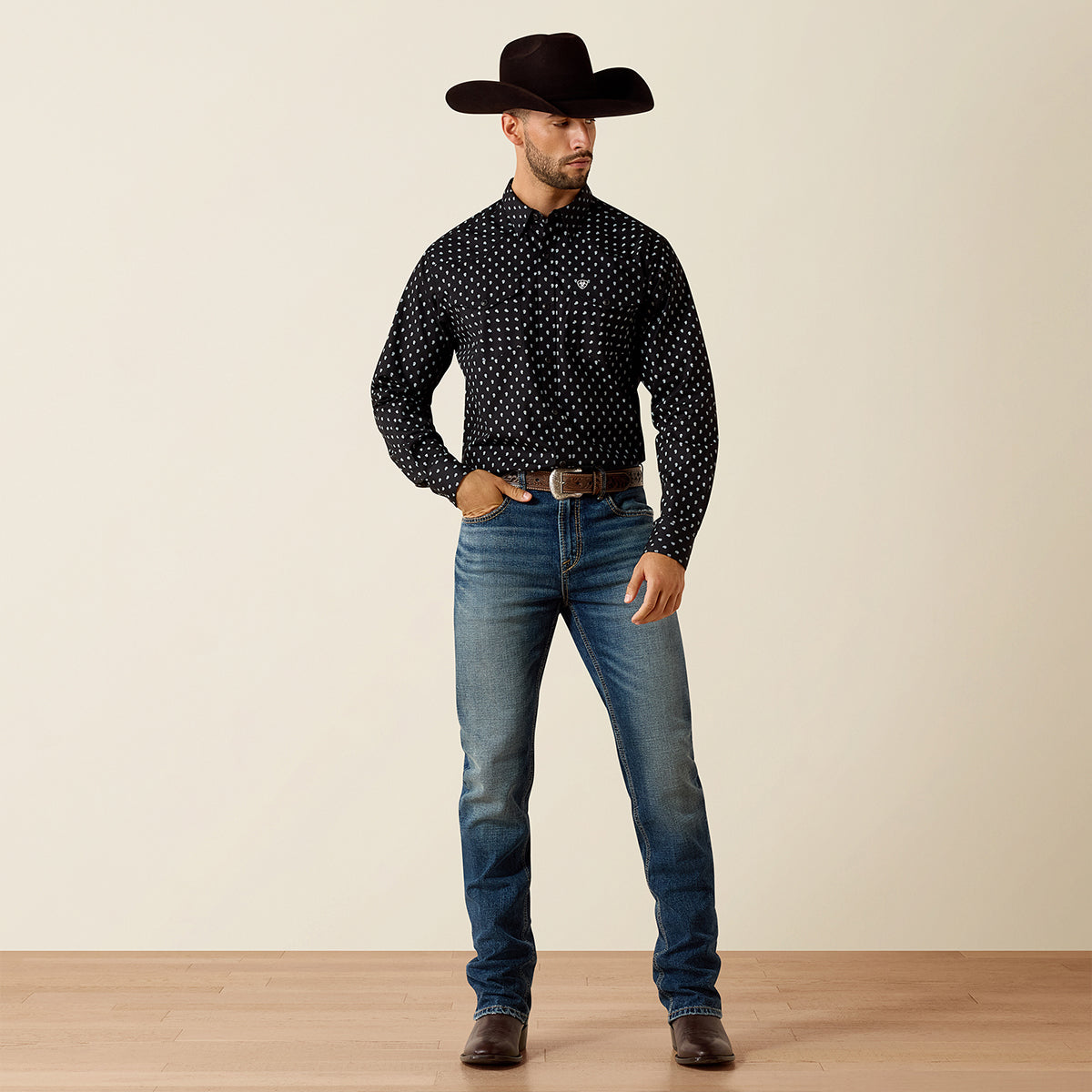Full-body view of a man wearing the Ariat Men's Black Paisley Truth Fitted long sleeve Western snap shirt paired with medium-wash blue jeans, a black cowboy hat, and dark brown boots.