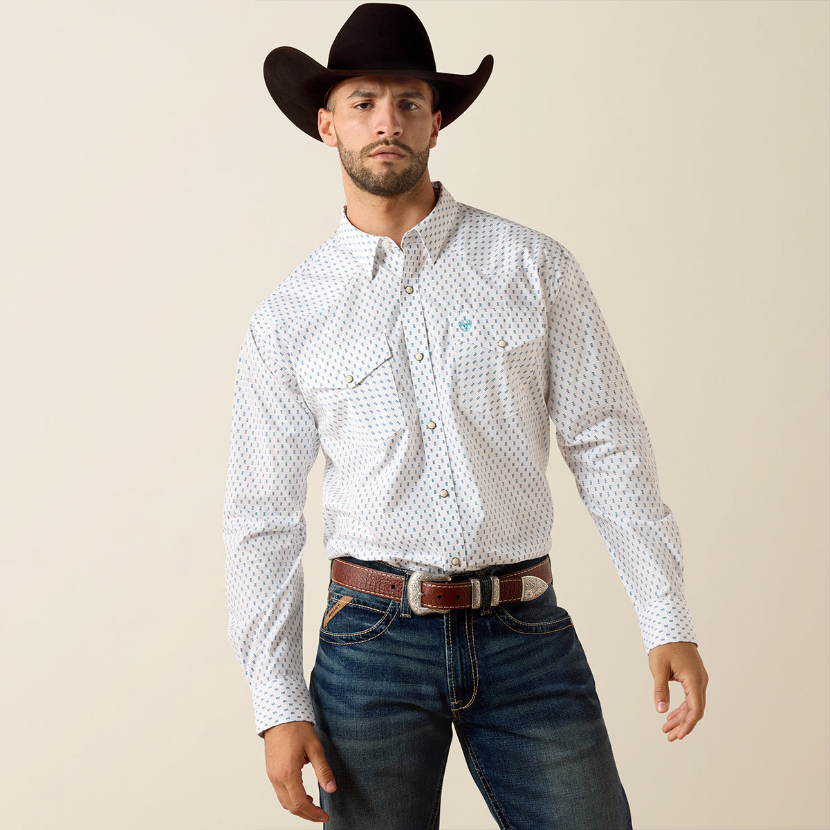 Man wearing a white patterned Ariat shirt, dark jeans, and a black cowboy hat against a beige background