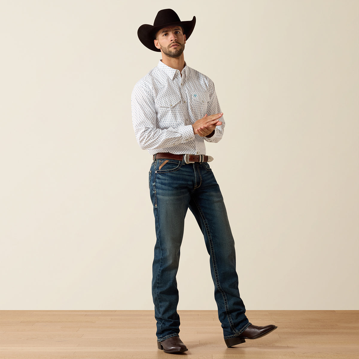 Man wearing a cowboy hat, patterned Ariat shirt, and jeans on a plain background