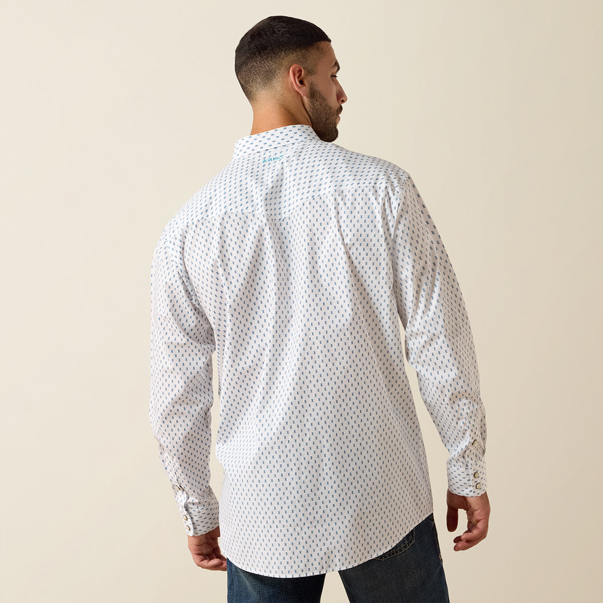 A man viewed from the back wearing a white long-sleeve button-up Ariat shirt with a small repeating blue and red geometric pattern, paired with dark blue jeans, against a solid beige background.