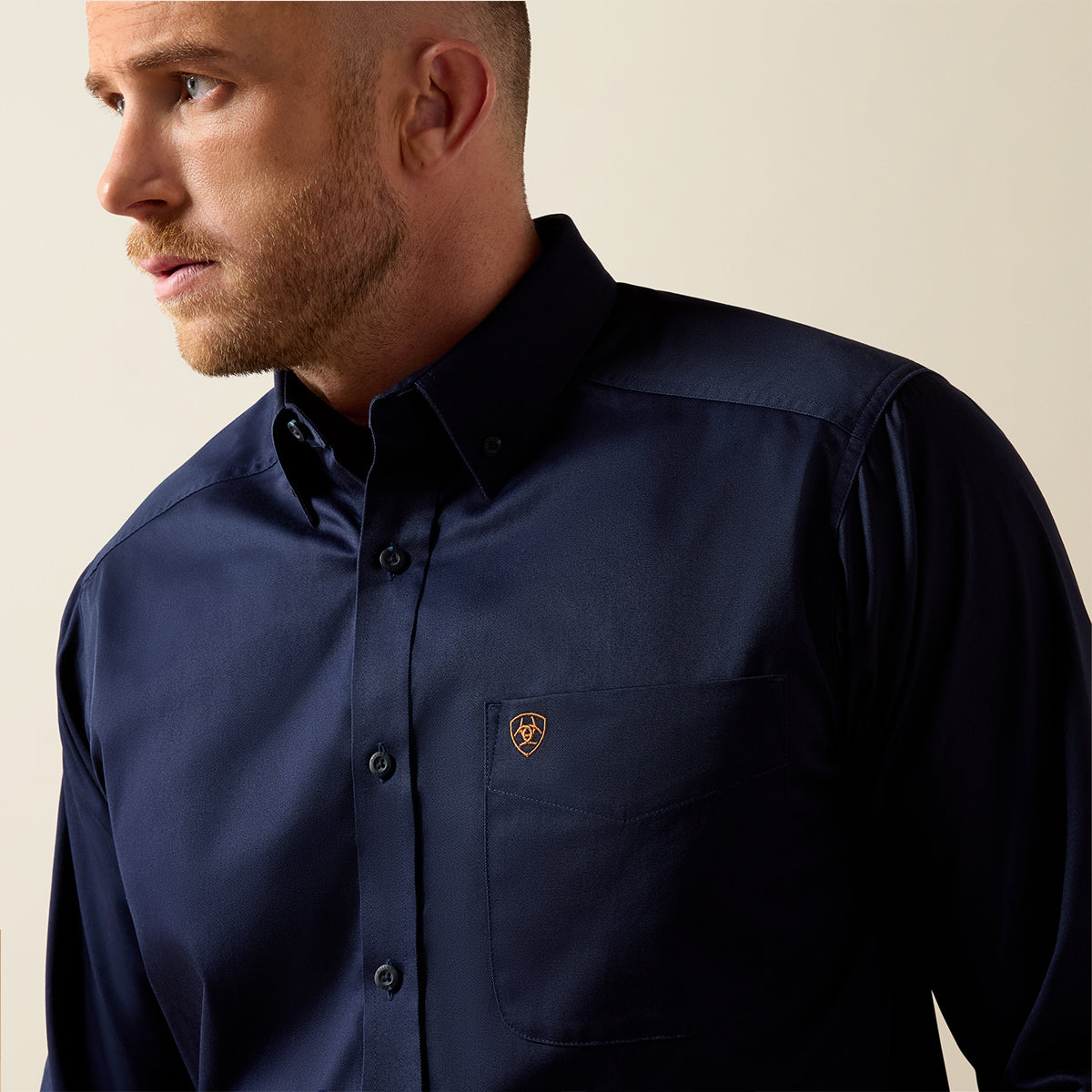A man wearing a dark navy blue button-down shirt with a small shield logo on the chest pocket, photographed against a plain beige background.