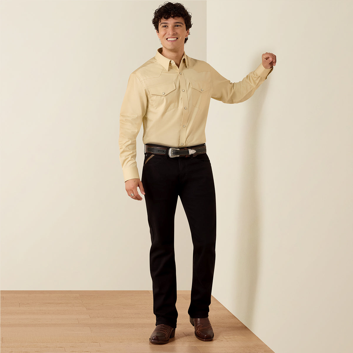 A man wearing an Ariat light beige long-sleeve western-style button-down shirt, black pants, a black belt, and brown shoes, standing against a cream-colored wall on a light wood floor.