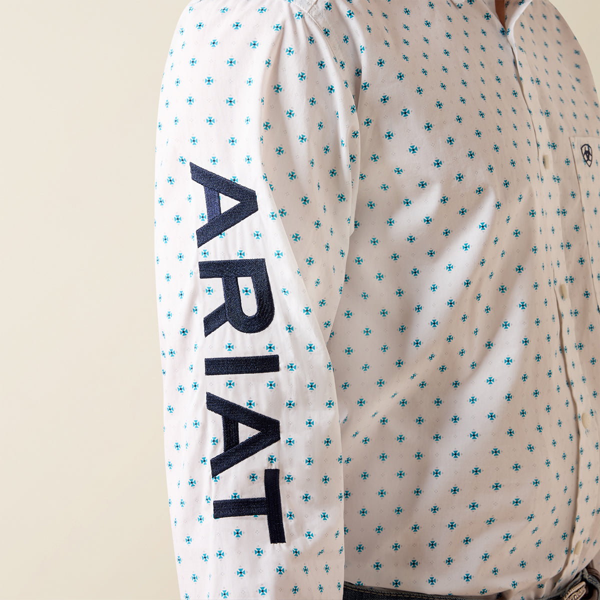 Close-up of a white long-sleeve button-up shirt with a blue geometric pattern and large navy ARIAT text printed vertically down one sleeve.