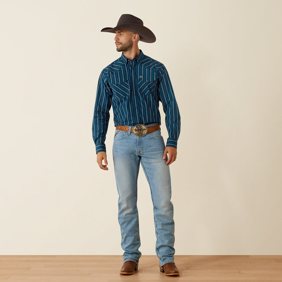 A man wearing a dark brown cowboy hat, a blue Ariat vertically striped western shirt, light blue jeans, and brown leather boots, standing against a plain beige studio background.