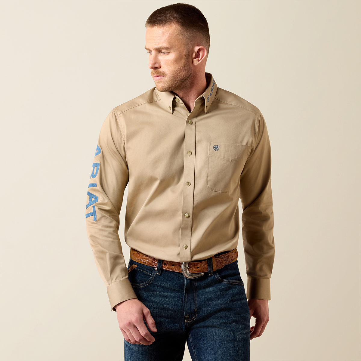 Front and full-body of a man wearing the Ariat Men's Team Logo Twill Savannah Tan long sleeve button-down shirt with navy embroidery, jeans.