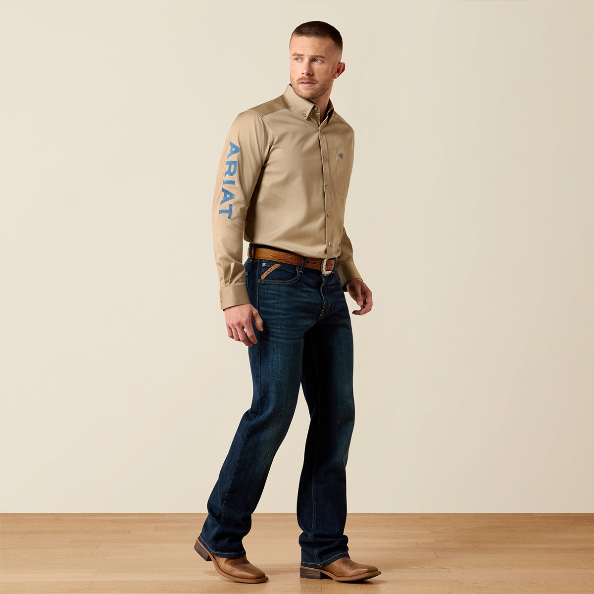 A man modeling a beige Ariat long-sleeve button-down shirt with blue vertical branding on the sleeve, paired with dark blue jeans and brown leather shoes, against a plain beige studio background.