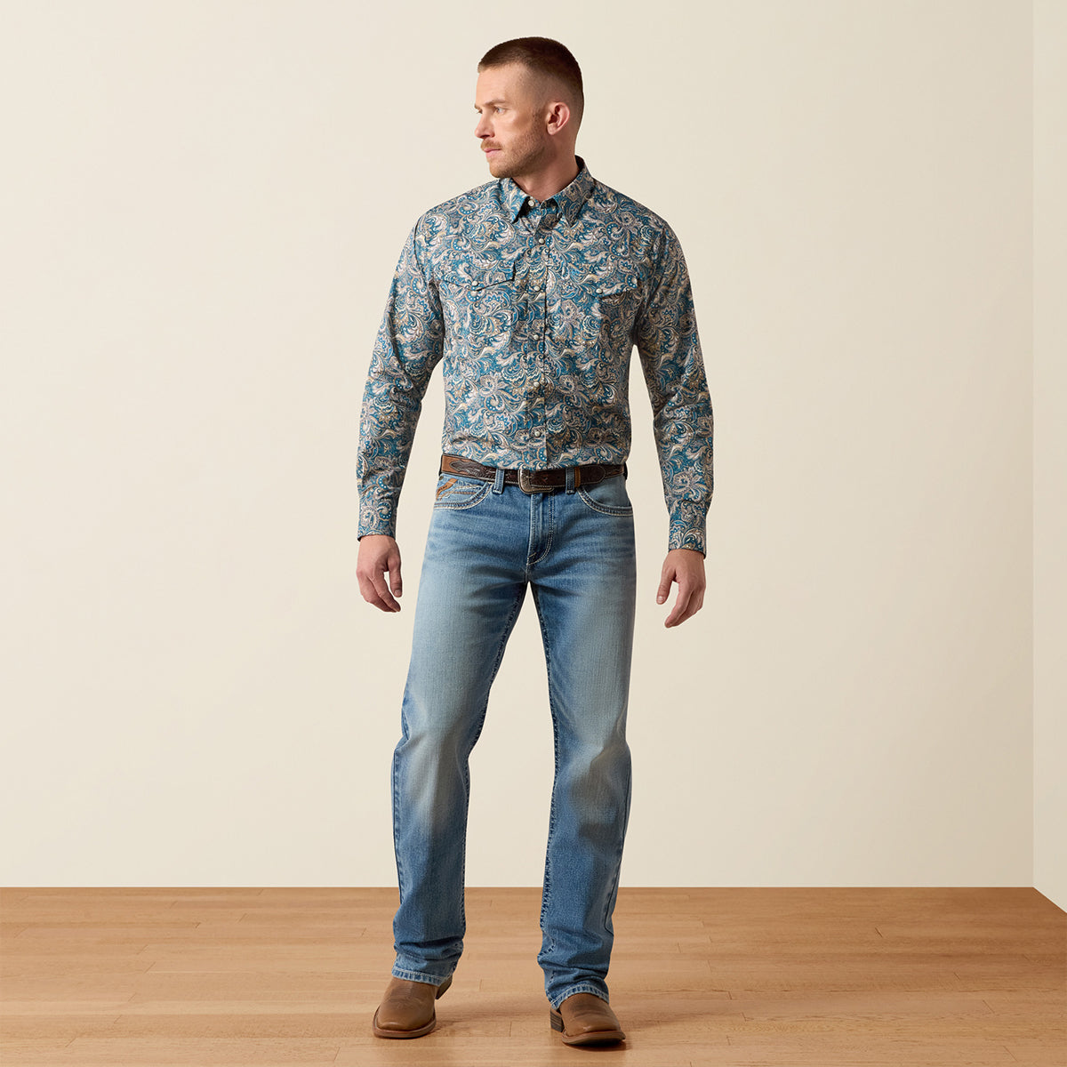 A man wearing a long-sleeve button-down Ariat shirt with a blue and beige paisley pattern, paired with light blue jeans and brown shoes, standing against a plain beige wall and light wood floor.