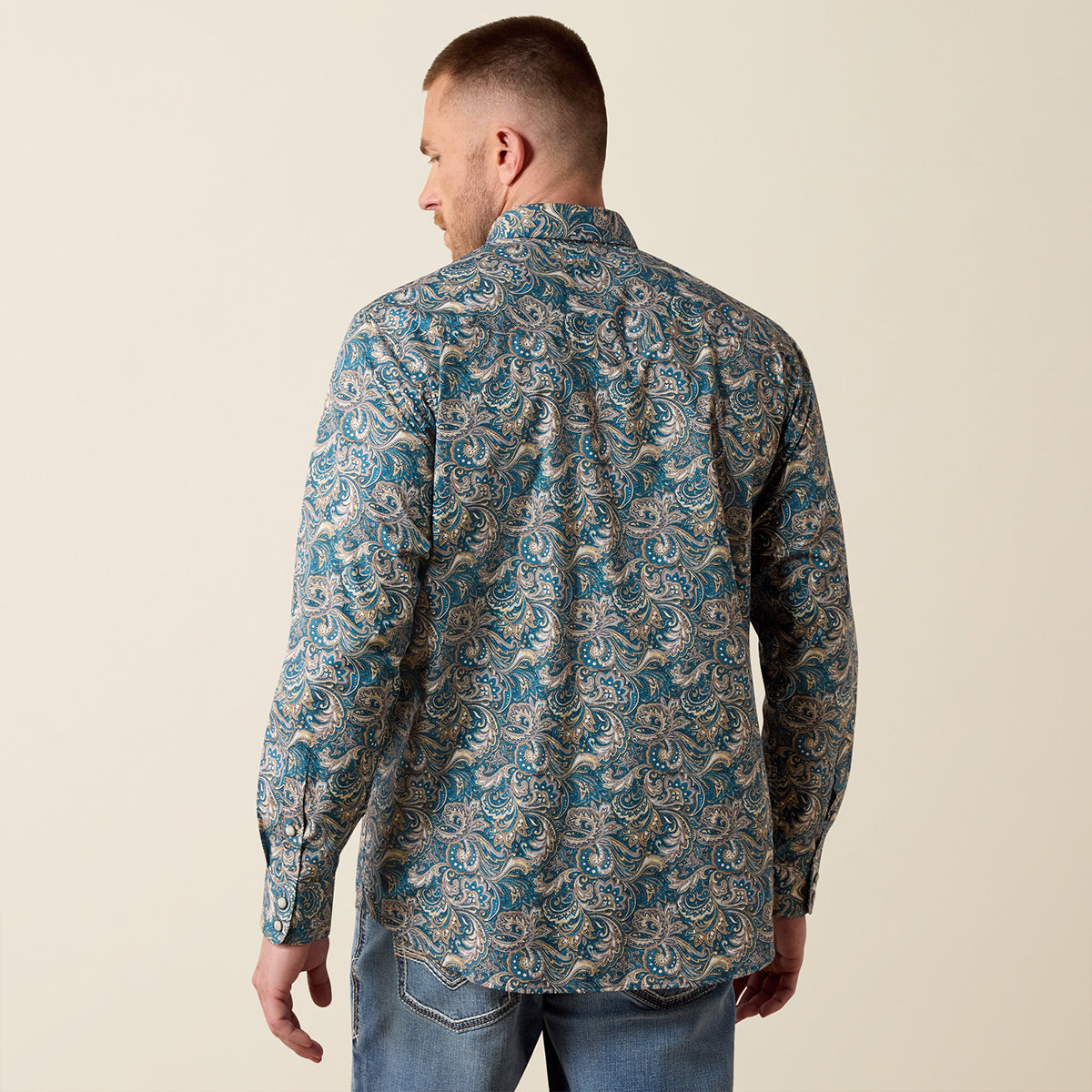 A man viewed from behind wearing a long-sleeve button-up shirt with a blue, beige, and gray paisley pattern, paired with blue jeans, against a solid beige background.