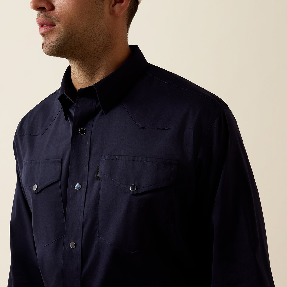 A close-up of a man wearing a dark navy western-style button-down shirt with two chest pockets and silver-toned snap buttons, set against a plain beige background.