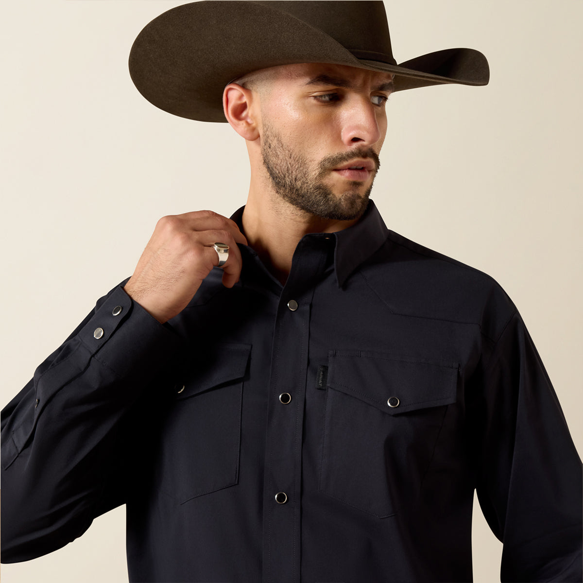 A man wearing a black western-style long-sleeve button-up shirt and a brown cowboy hat, shown from the chest up against a plain light beige studio background.
