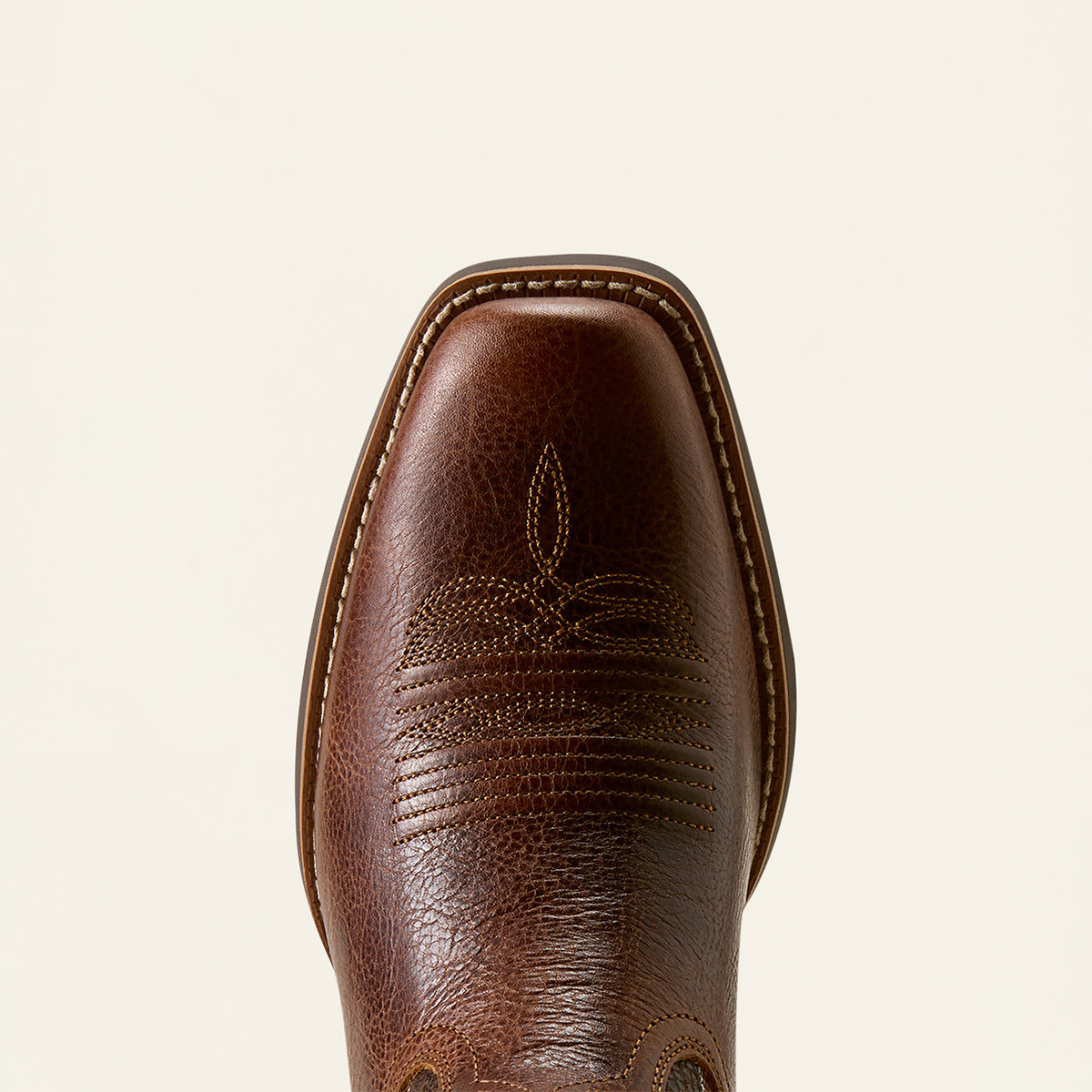 A close-up top-down view of a brown leather Ariat boot square toe, showing detailed stitching, a decorative floral tooling pattern, and a square toe shape against a light cream background.