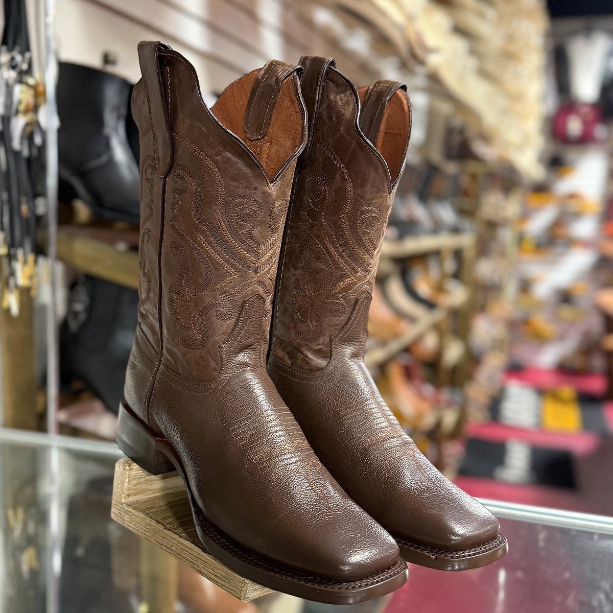 A pair of brown leather cowboy boots with decorative stitching, displayed on a wooden stand inside a western store.