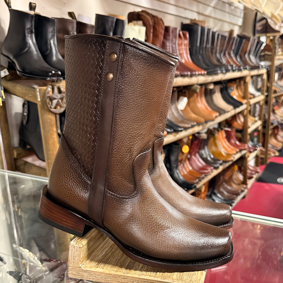 A pair of brown textured leather mid-calf boots displayed on a wooden stand inside a boot shop, with shelves of other boots blurred in the background.