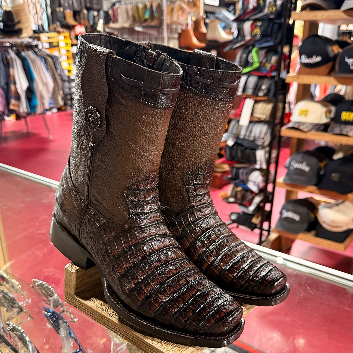 A pair of dark brown crocodile print cowboy boots with semi square toe and a decorative snap detail on the back, displayed on a wooden stand inside a western store.