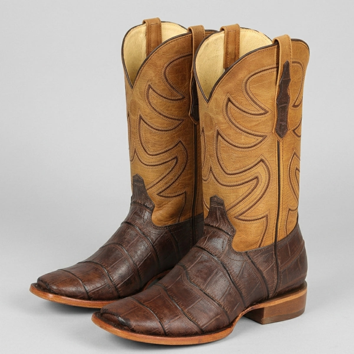 Pair of men's Alligator print brown and tan square toe cowboy boots on a light gray background.