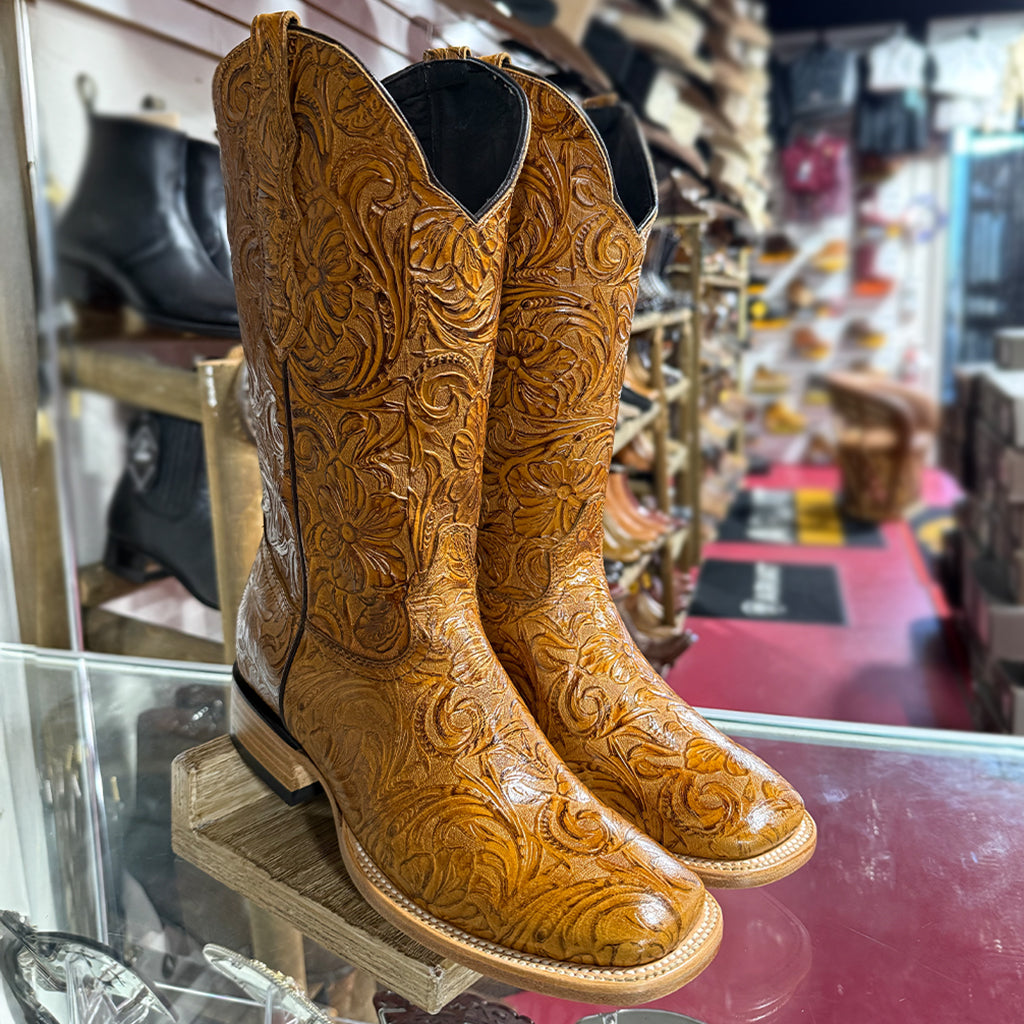 A pair of women's tall tan cowgirl boots featuring an intricate floral tooled leather print pattern, a wide square toe, and double-stitched leather welts.