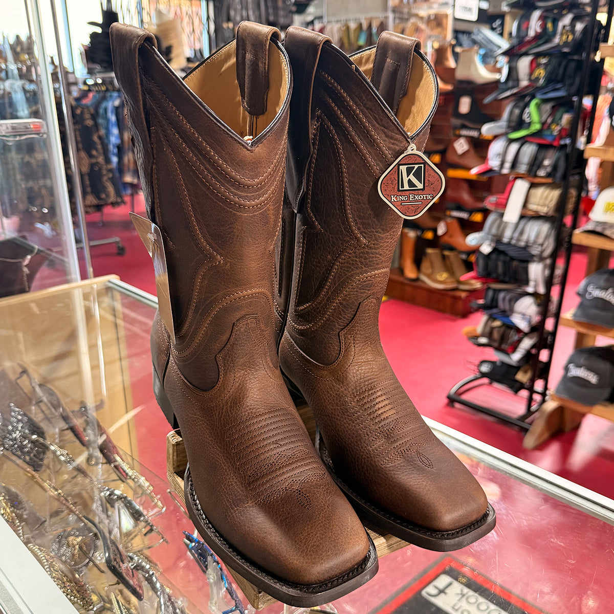King Exotic men's brown leather cowboy boots displayed on a glass counter inside a retail store. The boots feature decorative stitching, a square toe.