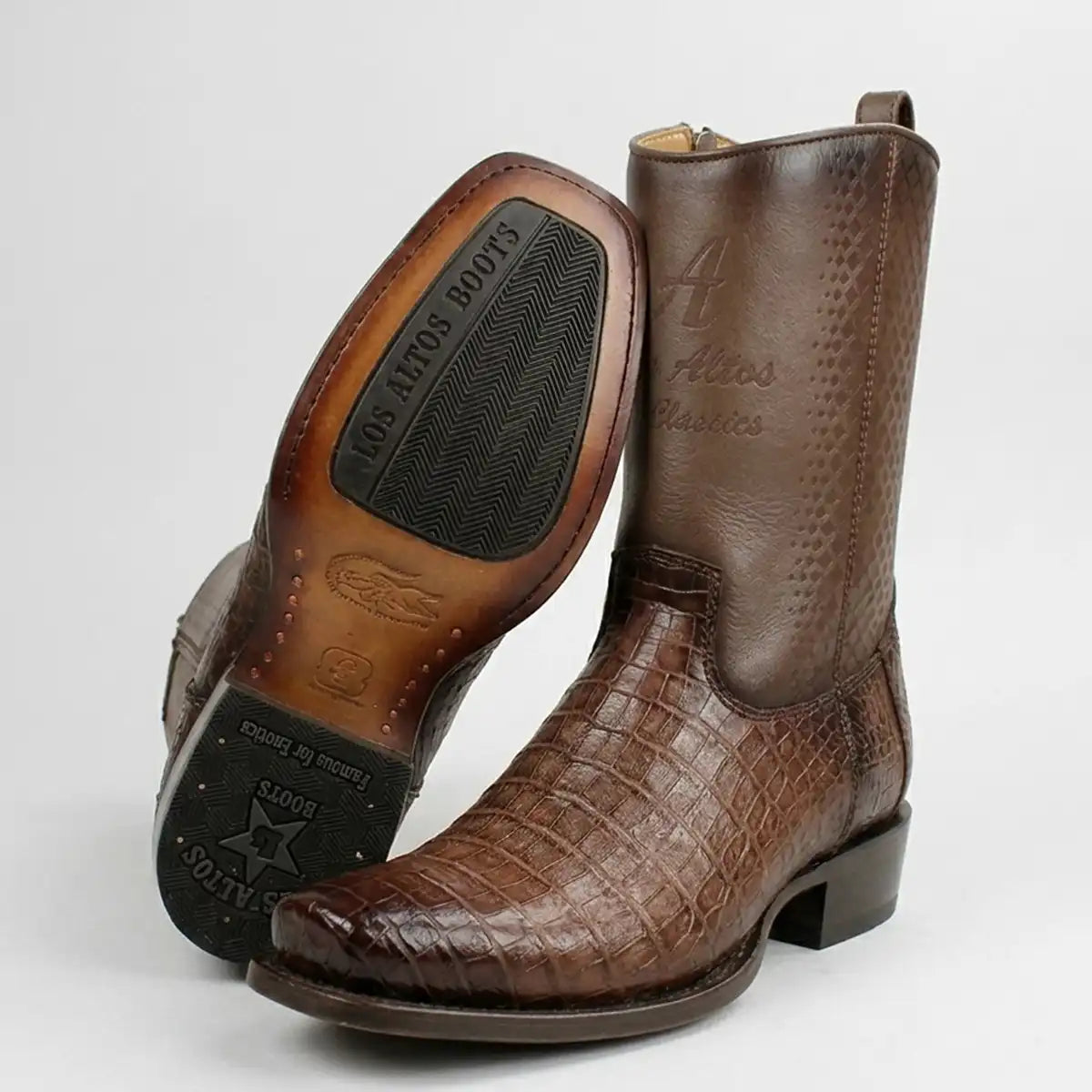 A pair of brown caiman leather square toe western boots with, shown on a light gray background. One boot is standing upright to show the front and heel, while the other is turned on its side to display the sole, which features the brand name 'Los Altos Boots'.