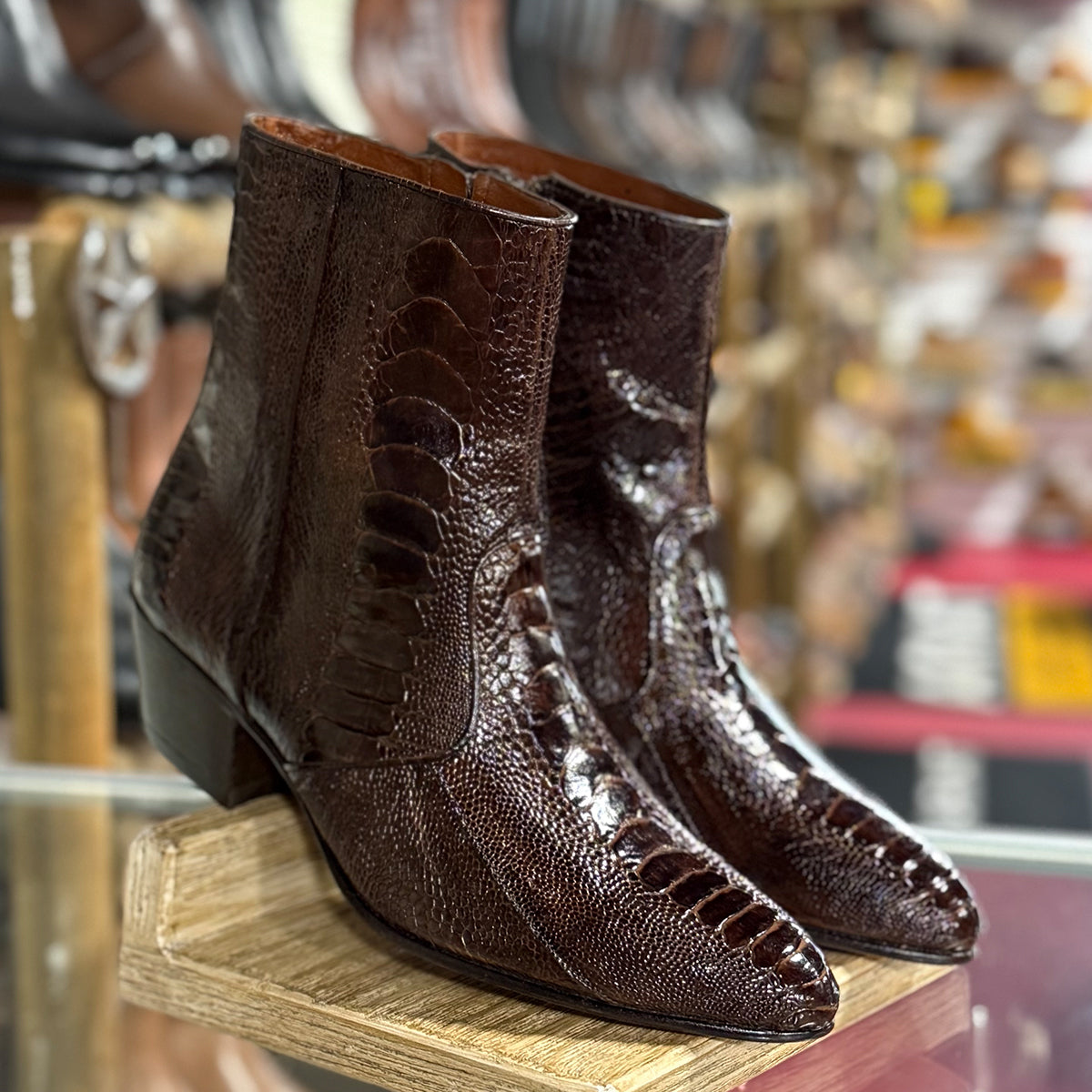 A pair of Los Altos men's dark brown ostrich leg ankle boots with pointed toes and low block heels, displayed on a wooden stand in a western store setting.