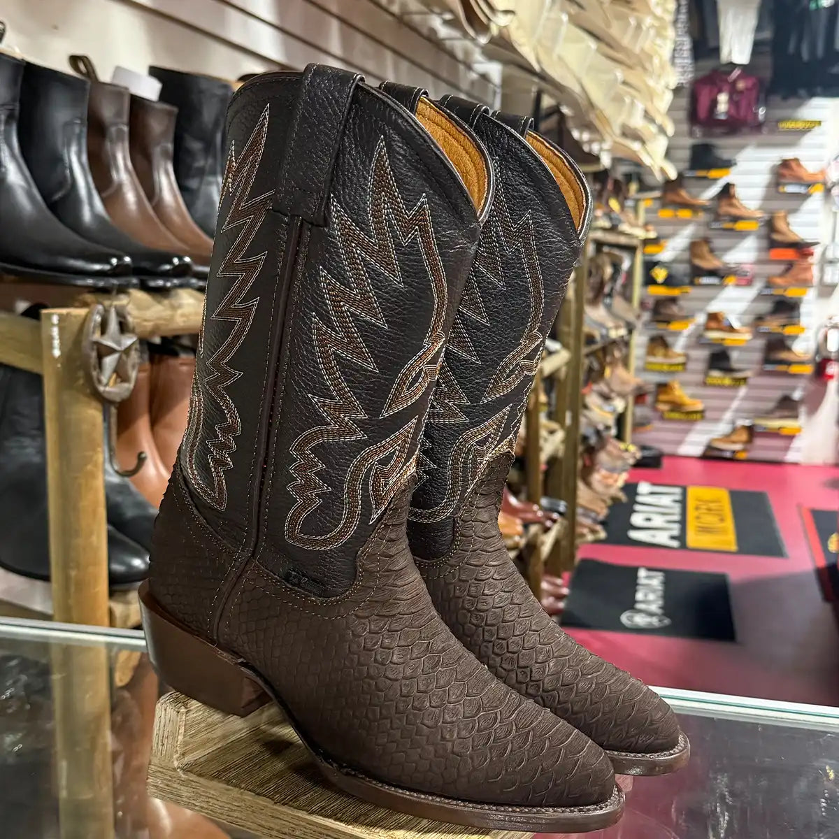 A pair of dark brown python print cowboy boots with embroidered western stitching, displayed on a glass shelf inside a western store.