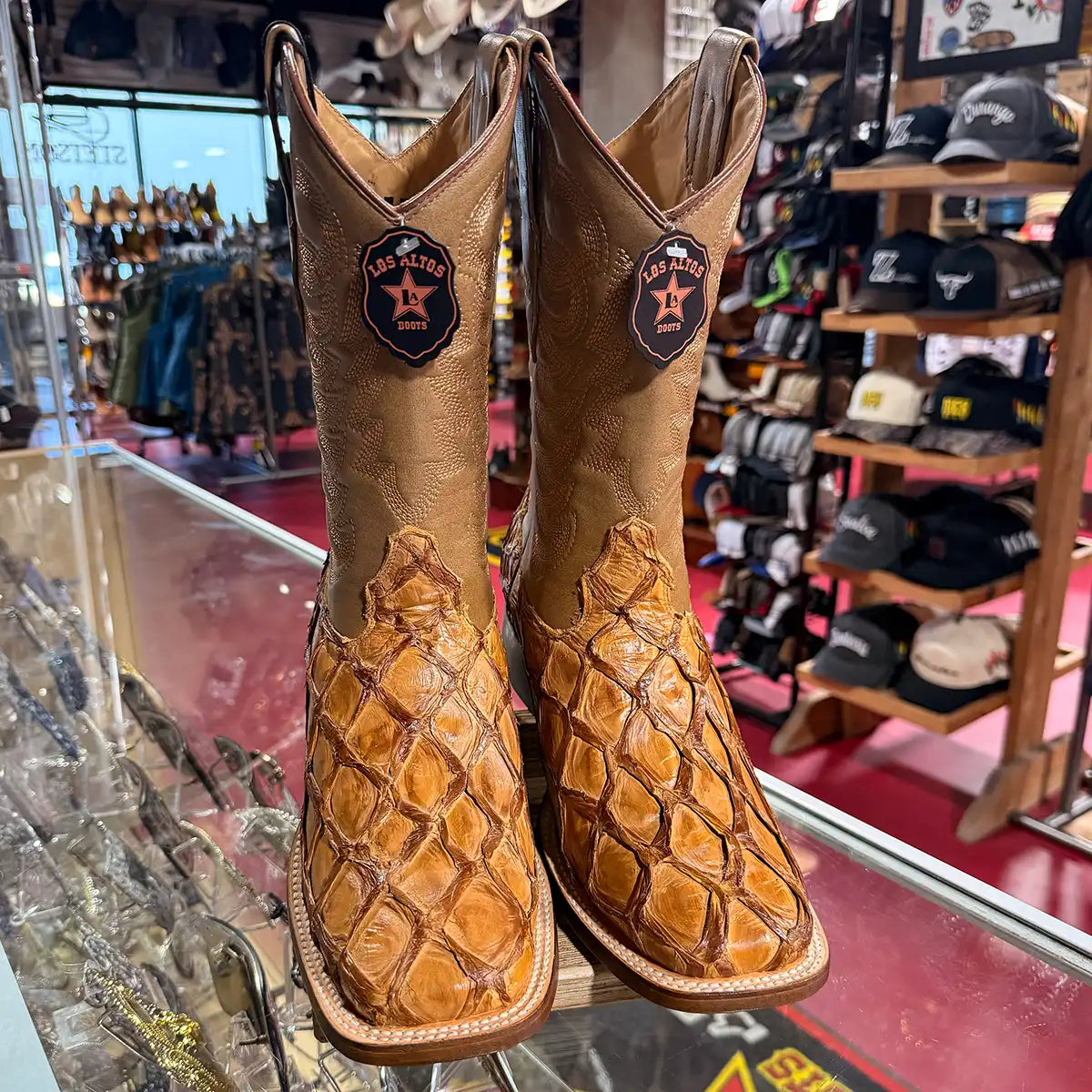 A pair of Los Altos Pirarucu tan cowboy boots with a stitched shaft, displayed on a glass case inside a western store.