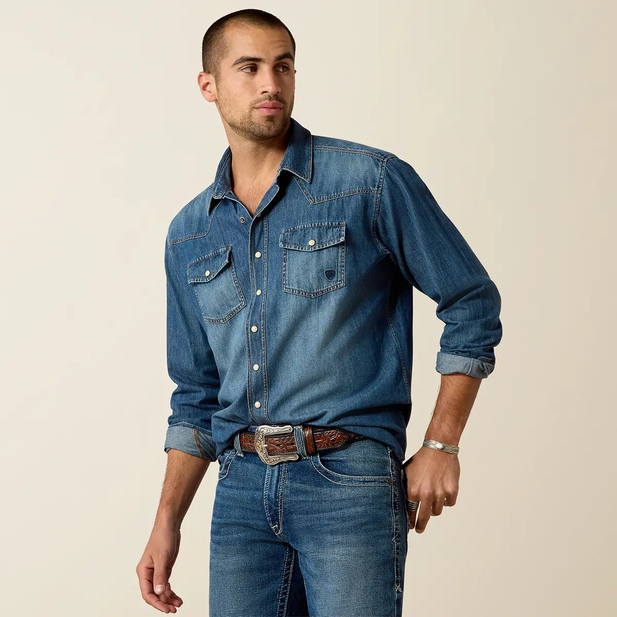 Medium shot of a model wearing the Ariat Men's Stone Blue Denim Retro Fit shirt tucked into blue jeans with a Western silver buckle belt.