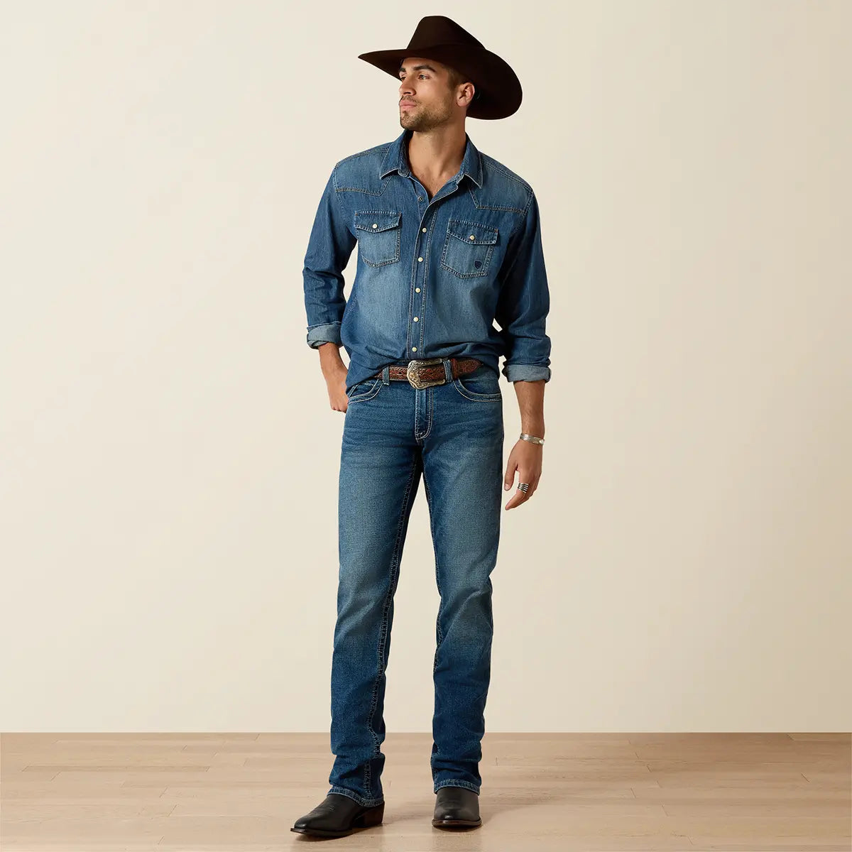 Full-length lifestyle shot of the model wearing the Ariat Stone Blue denim shirt, dark cowboy hat, leather belt, and dark Western boots.