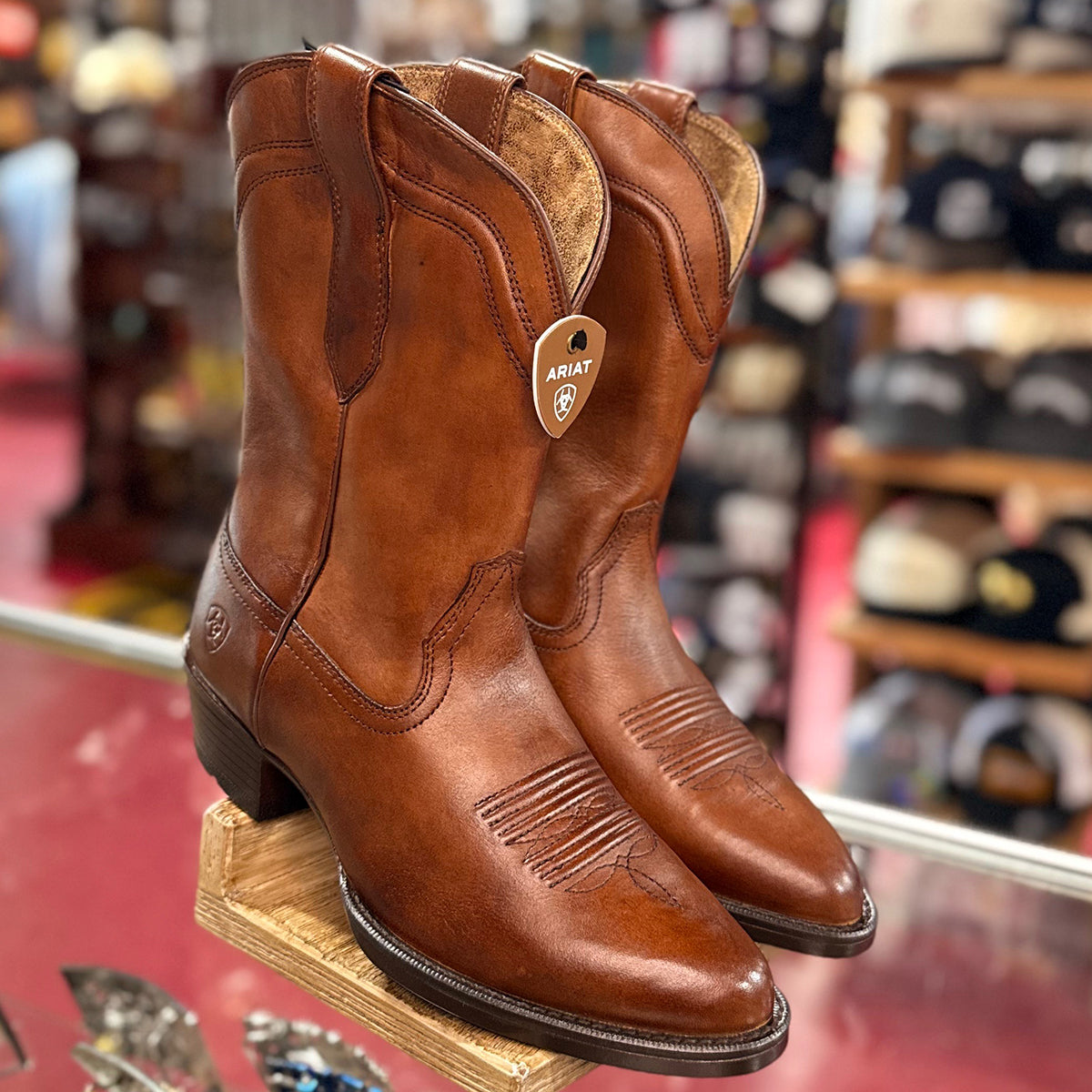 Pair of men's brown Ariat cowboy boots on a wooden block with a store background.