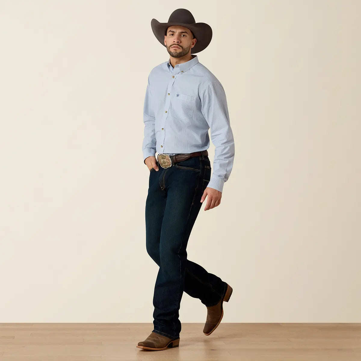 Full-length lifestyle shot of a model walking in the Ariat Pro Series Dayne True Blue shirt, dark cowboy hat, and Western leather boots.