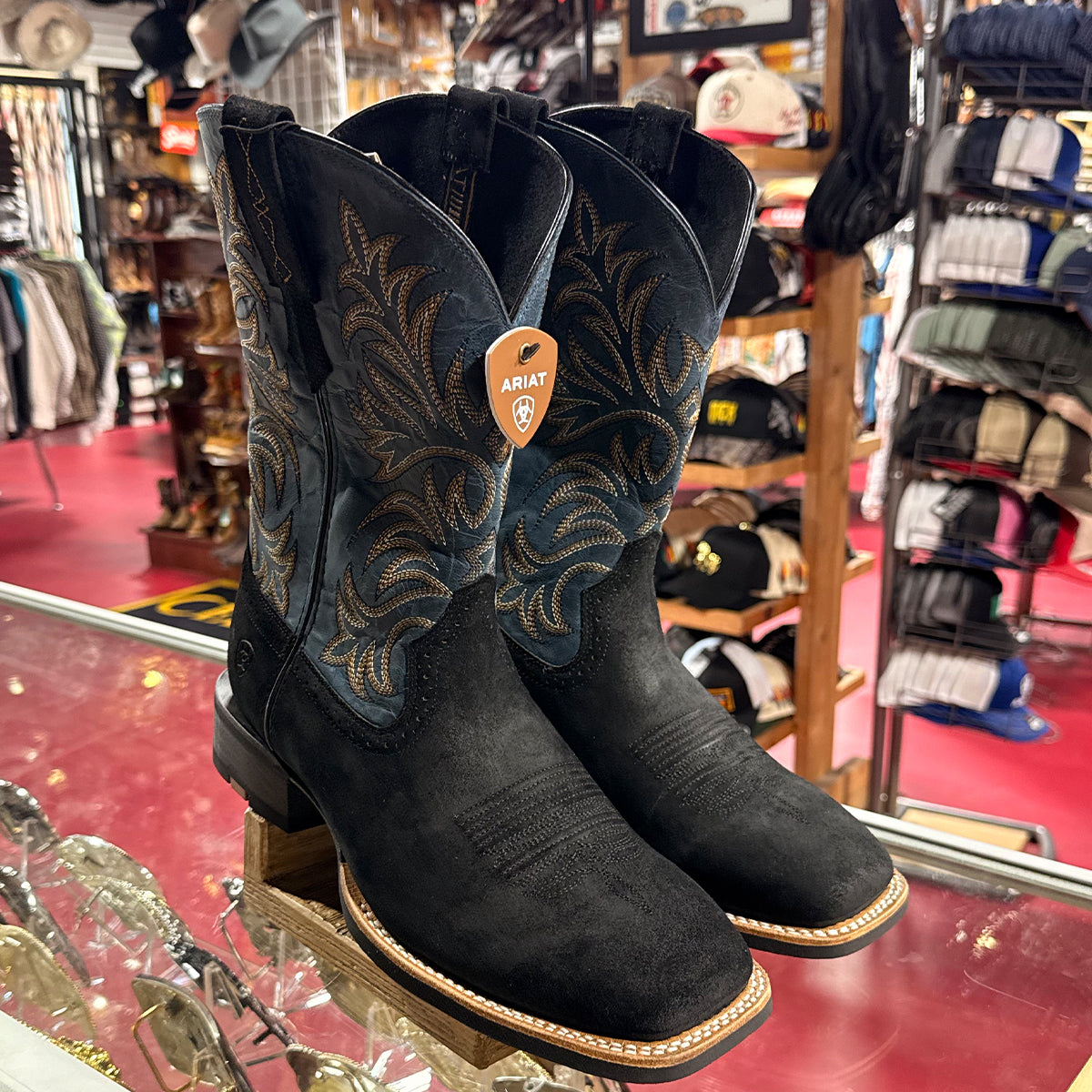 Black suede Ariat boots on display in a store with various items in the background.