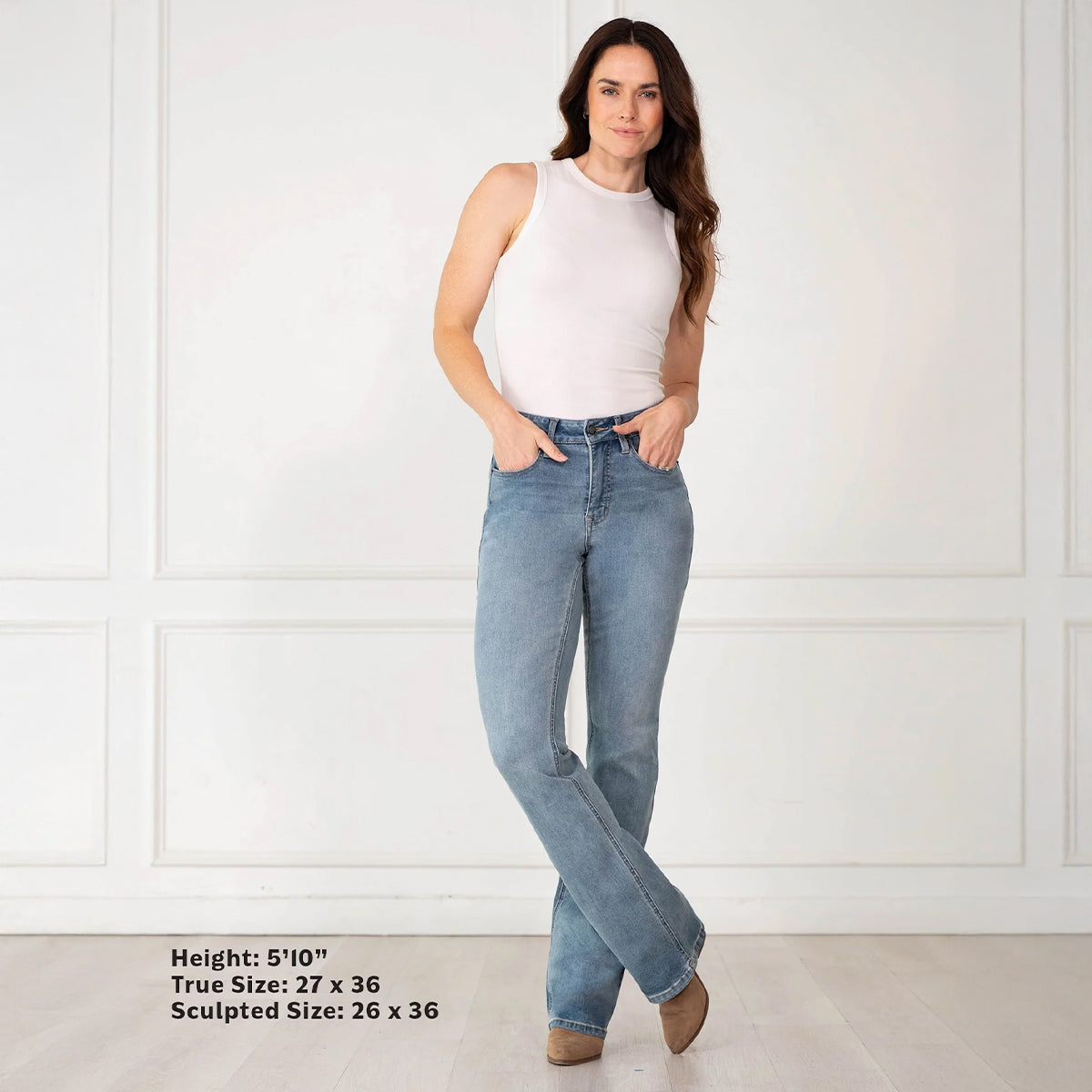 A woman modeling light-wash blue jeans with a white sleeveless top, standing against a white paneled wall. Text at the bottom left lists height and true/sculpted sizes.