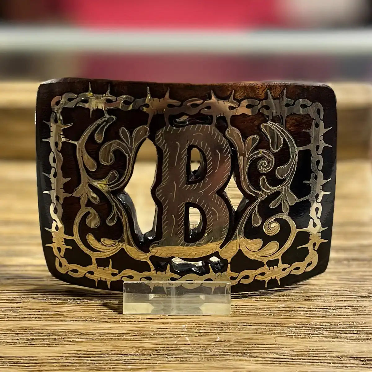 Front view of the 4 1/4" x 3" handcrafted mesquite wood belt buckle showing the metal Monogram B inlay and barbed wire frame displayed on an acrylic stand.