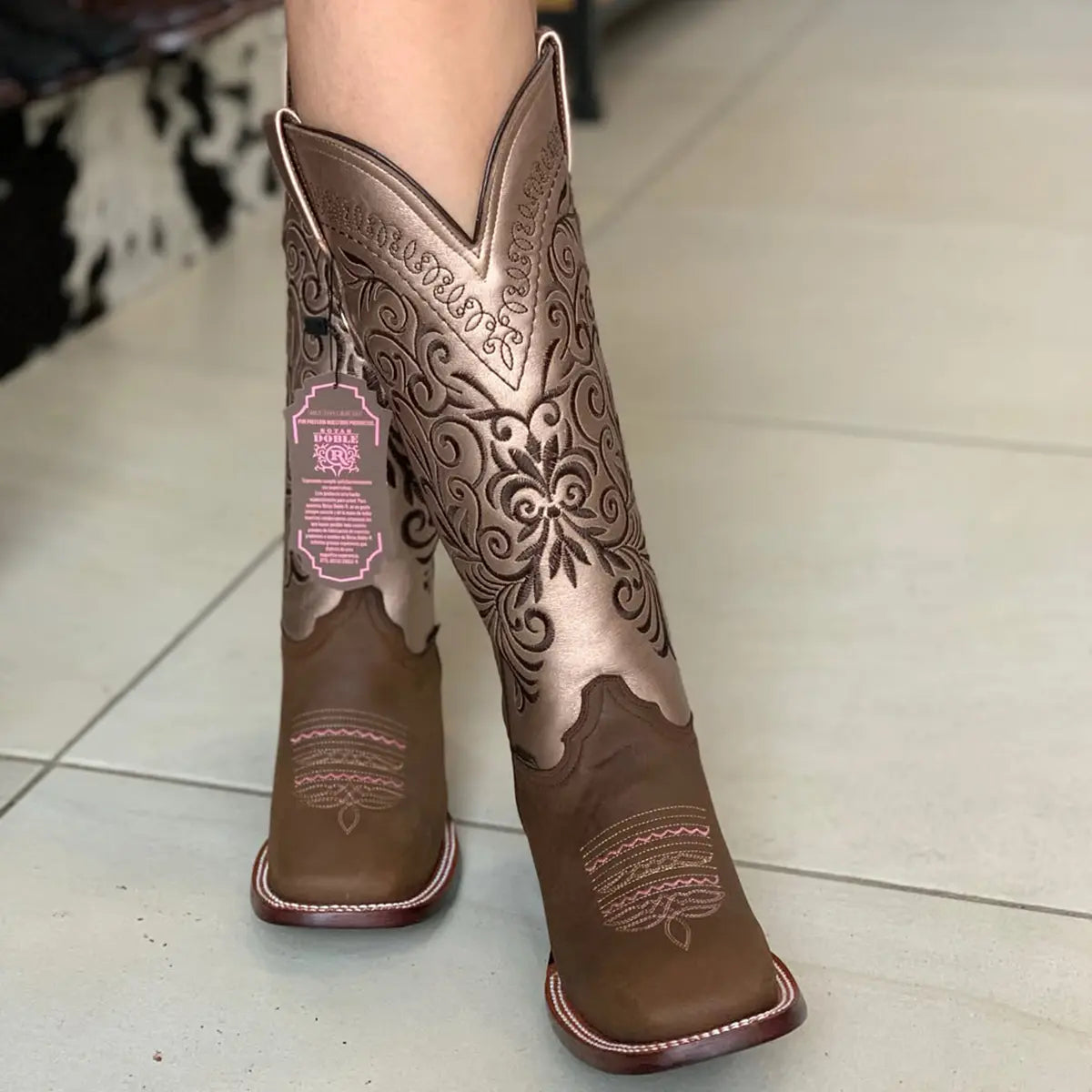 Front view of a model wearing Botas Doble R Ivanna Crazy Cafe boots, emphasizing the square toe profile, pink vamp stitching, and the metallic deep V-cut shaft with a pink brand tag attached.