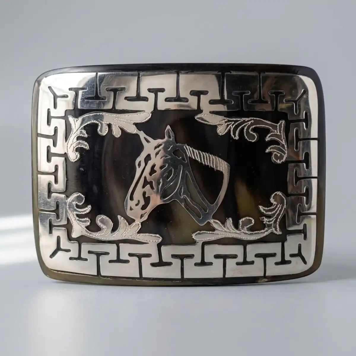 A rectangular silver-toned belt western buckle featuring an engraved horse head motif in the center, surrounded by decorative scrollwork and a Greek key border pattern, set against a dark tortoiseshell background.