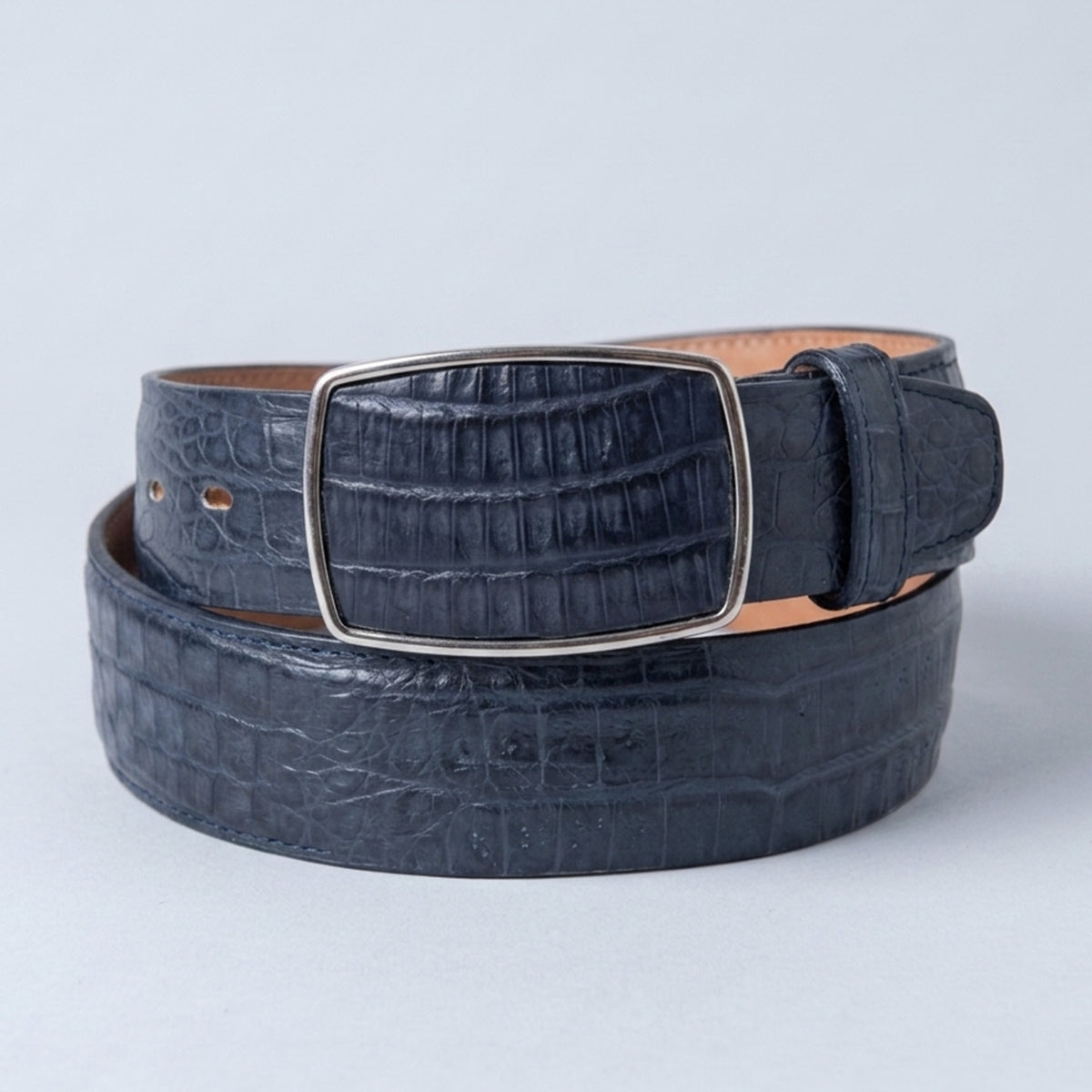 A dark navy blue caiman belly leather belt with a silver-tone rectangular buckle, coiled on a light gray background.