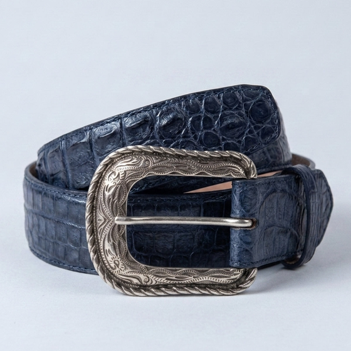 A dark navy blue crocodile hornback leather belt with a  silver-toned western-style buckle, coiled on a light gray background.