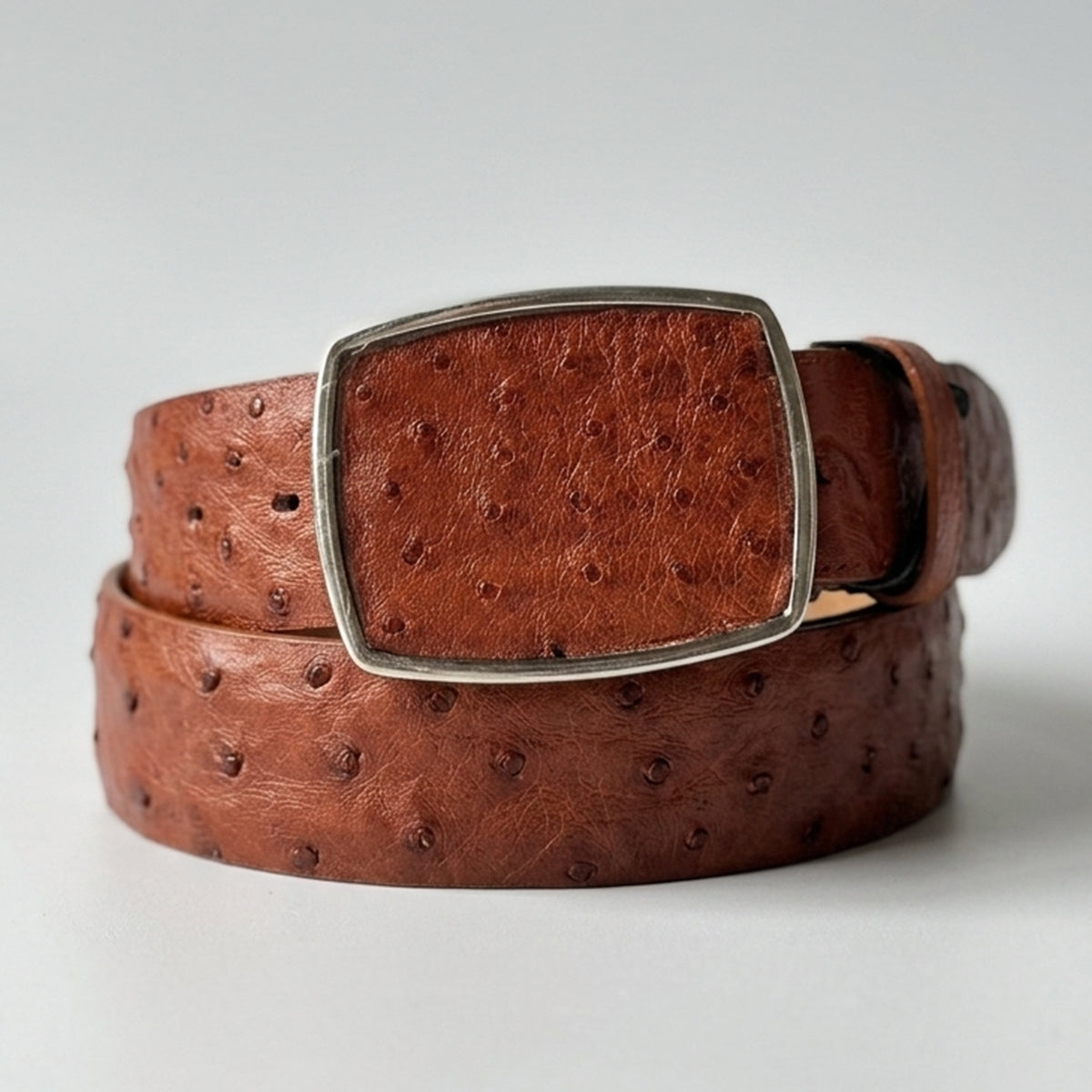Los Altos men's cognac brown ostrich leather belt with a silver-tone rectangular buckle, coiled on a light gray background.